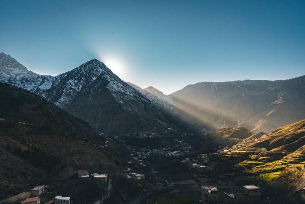 A view of sunset sunrise with sunstar in rural mountain village Imlil in High Atlas mountains Morocco in Africa. Snow covered peaks in background with lightrays and blue sky. - Fotó, kép