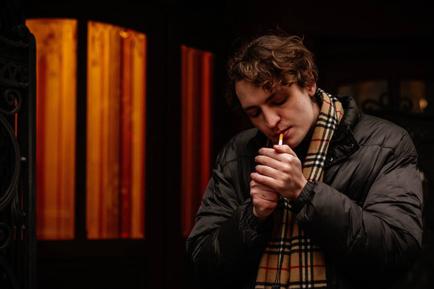Όμορφος κομψός νέος χίπστερ άντρας καπνίζει τσιγάρο στο δρόμο το χειμώνα, κοιτάζοντας αλλού. Πορτρέτο του όμορφου άντρα ντυμένο άνετα. Vintage ευρωπαϊκή πόλη, φώτα στο παρασκήνιο. Μαλακή εστίαση. - Φωτογραφία, εικόνα