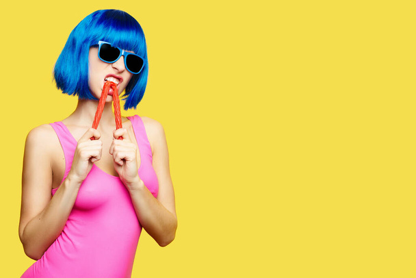 Mavi peruklu güneş gözlüklü ve pembe mayo giyen kız sarı arka planda lolipop yiyor. - Fotoğraf, Görsel