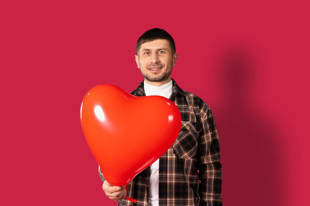 A guy holds out to the camera a red heart-shaped balloon on a red background. St.Valentine's Day. High quality photo - Φωτογραφία, εικόνα