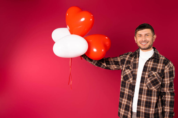 Portrait of a smiling man holding up in air a bunch of red balloons over red background with empty space. St.Valentine's Day. High quality photo - Φωτογραφία, εικόνα