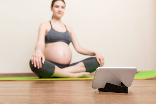 Pregnant female holding smartphone while sitting on exercise yoga mat at home. Fitness or yoga training online at coronavirus time. - Fotografie, Obrázek