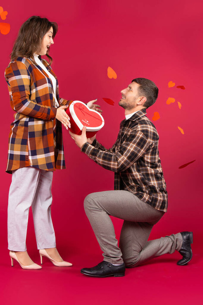 A man kneels in front of a surprised girl and gives a heart-shaped gift box. Awesome background and red hearts. High quality photo - Valokuva, kuva