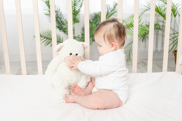 baby six months playing in a crib in a white bodysuit with a Teddy bear - Foto, Imagen