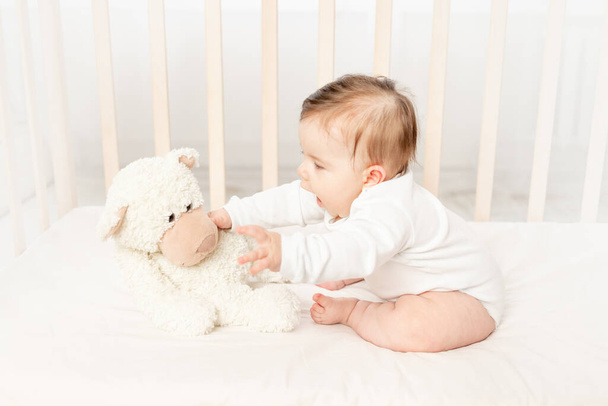 bebé de seis meses jugando en una cuna en un body blanco con un osito de peluche - Foto, imagen