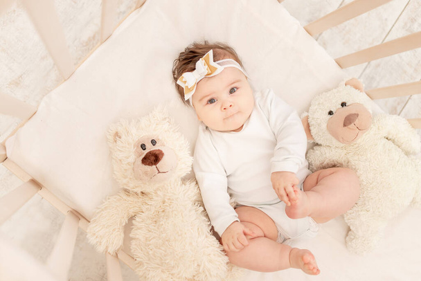 o bebê tem seis meses de idade em um berço em um terno branco com um ursinho de pelúcia - Foto, Imagem