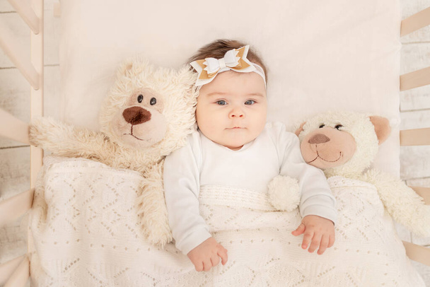 de baby is zes maanden oud in een wieg in een wit pak met een teddybeer - Foto, afbeelding