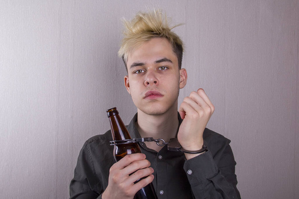 A teenager in handcuffs and with a bottle in his hand on a gray background. Concept: adolescent alcoholism, early attachment to drinking, poor heredity. - Photo, Image