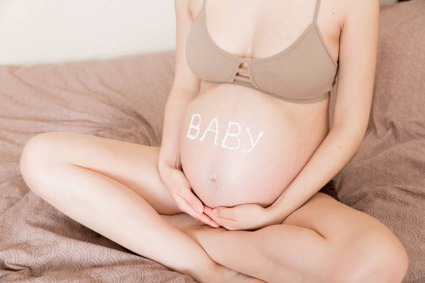 een zwanger meisje zit thuis op het bed en smeert een anti-stretch crème op haar buik. Zwangerschap, moederschap, voorbereiding en verwachting concept.het woord baby op de buik. - Foto, afbeelding