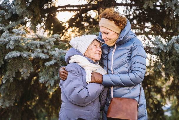 Concept of happy family, old age, emotions, senior care in retirement age. Active senior grandmother and adult daughter hugging outside in winter against background of Christmas tree in forest. - Photo, Image