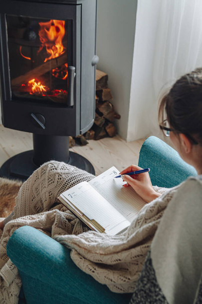 Young woman write in notebook sitting in armchair by fireplace - Foto, imagen