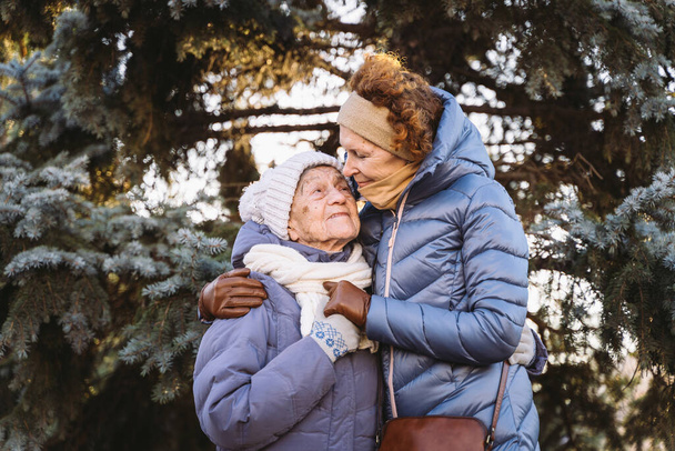 Kaukaskie kobiety starsza matka i dojrzała córka w ogrodzie zimowym na tle jodły przytulają się twarzą w twarz, uśmiechają się, spędzają czas ze starymi rodzicami, spacerują ze starszą mamą w lesie na Boże Narodzenie - Zdjęcie, obraz
