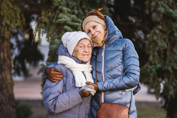 Caucasian women senior mother and mature daughter in winter garden on background fir tree hug each other face to face, smile, spend time with old parents, walk with elderly mom in forest for Christmas - Photo, Image