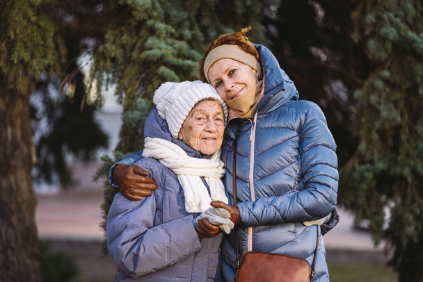 Pojęcie szczęśliwej rodziny, starości, emocji, opieki seniorskiej w wieku emerytalnym. Aktywny starszy babcia i dorosły córka przytulanie na zewnątrz w zimie na tle choinki w lesie. - Zdjęcie, obraz
