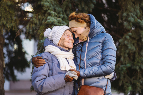 Concept van gelukkig gezin, ouderdom, emoties, ouderenzorg in de pensioengerechtigde leeftijd. Actieve grootmoeder en volwassen dochter knuffelen buiten in de winter tegen de achtergrond van kerstboom in het bos. - Foto, afbeelding