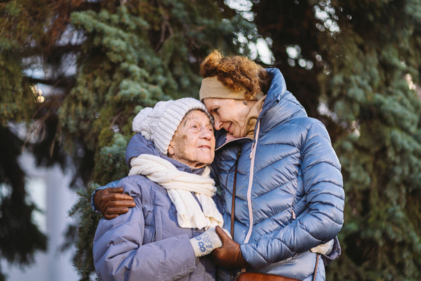 Las mujeres caucásicas madre mayor e hija madura en el jardín de invierno en el abeto de fondo se abrazan cara a cara, sonríen, pasan tiempo con los viejos padres, caminan con mamá anciana en el bosque para Navidad - Foto, imagen