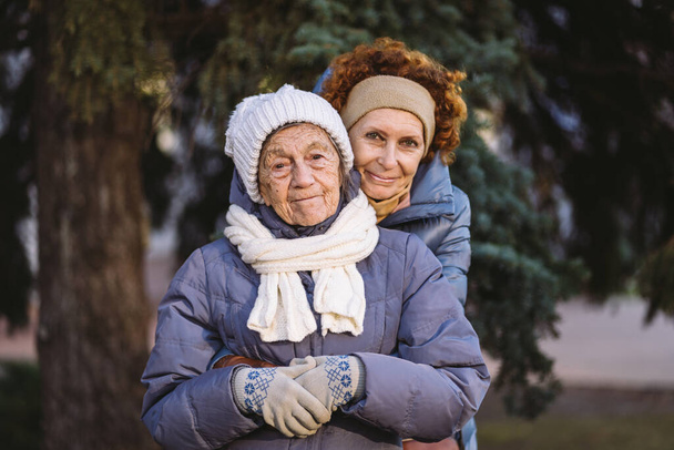 Caucasian women senior mother and mature daughter in winter garden on background fir tree hug each other face to face, smile, spend time with old parents, walk with elderly mom in forest for Christmas - Photo, Image