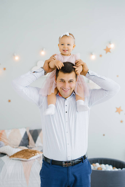 Happy father holds his one-year-old daughter around his neck in the children's room. They are happy - Photo, Image