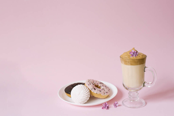 Kaunis dalgonkahvijuoma. Kuppi kahvia vaahto ja lila kukka, lautasellinen makeisia: vaahtokarkkeja ja donitseja lila tausta. Kopiointitila - Valokuva, kuva