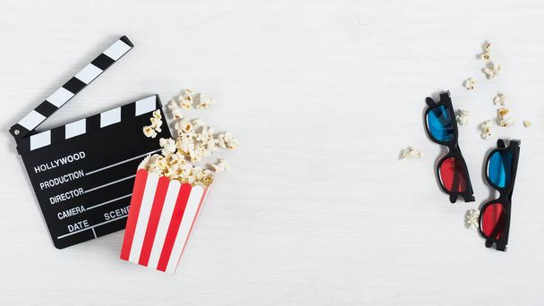Kino-Klappbrett, bunte 3D-Brille und rot gestreifter Eimer mit Popcorn auf weißem Holzhintergrund. Kinohintergrund. - Foto, Bild