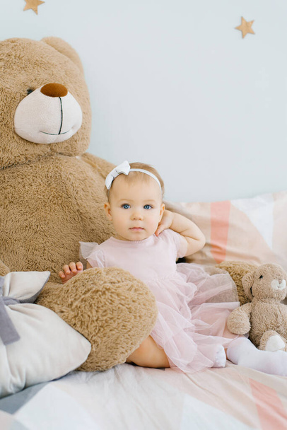 A cute one year old baby sits surrounded by soft animals toys and holds her hand on her neck - Fotografie, Obrázek