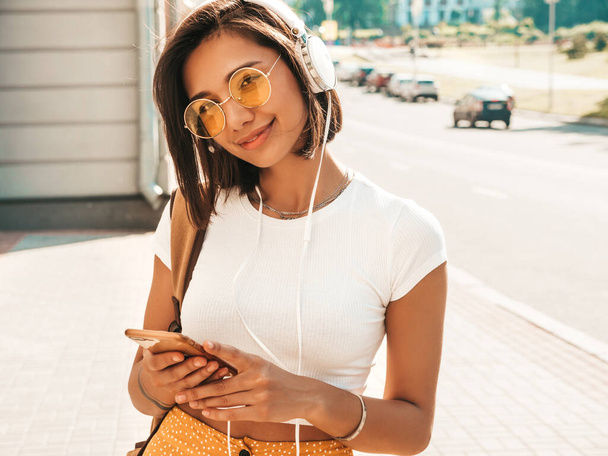 Fashion portrait of young stylish hipster woman walking in the street.Female wearing cute trendy outfit.Smiling model enjoy her weekends, travel with backpack. Listening to music via headphones - Фото, изображение