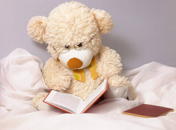Ένα χαριτωμένο μπεζ αρκουδάκι για να παίζουν τα παιδιά με γυαλιά διαβάζει ένα βιβλίο - Φωτογραφία, εικόνα