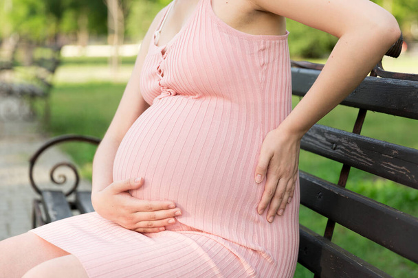 Eine schwangere junge Frau sitzt auf einer Bank im Park und hält sich am Bauch fest, der schmerzt. Schwangerschaft und Mutterschaft. Rückenschmerzen während der Schwangerschaft. - Foto, Bild