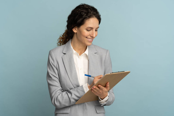 Teacher woman wear grey blazer and white blouse writing, makes notes in document, holds folder in her hand - Photo, Image