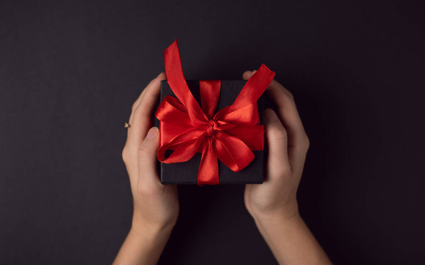 Woman's hand holding black gift box with red ribbon on black background. The Concept Of Valentine's Day. - Fotoğraf, Görsel