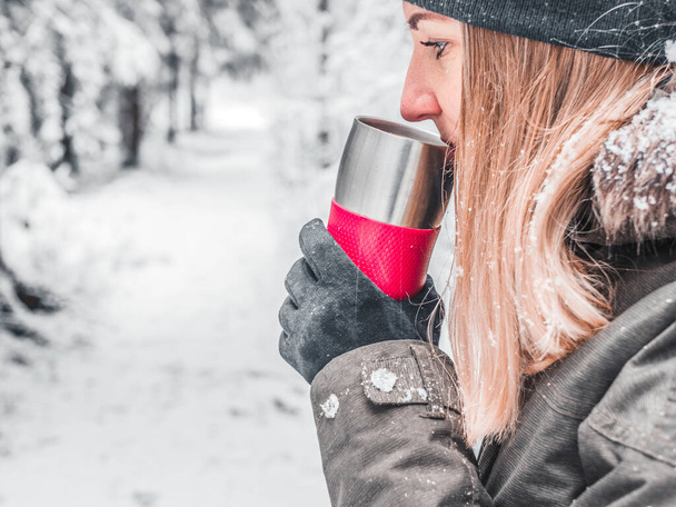 Mujer joven disfrutando de una bebida caliente en invierno. Bosque de abeto cubierto de nieve durante el invierno en el fondo. - Foto, imagen