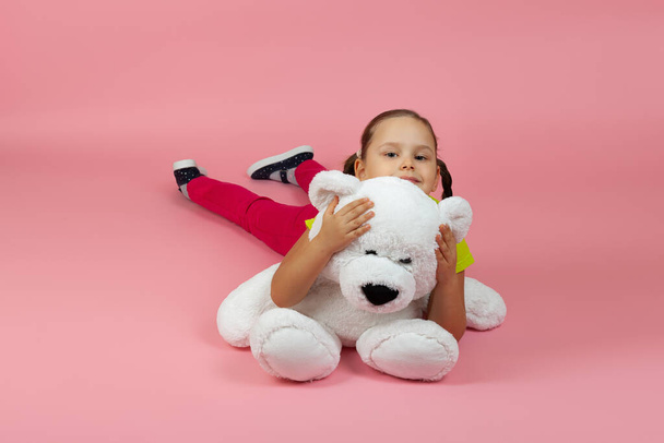 playful contented girl with ponytails and red pants and light green t-shirt lies on the floor and hugs white teddy bear isolated on a pink background - Foto, Imagen