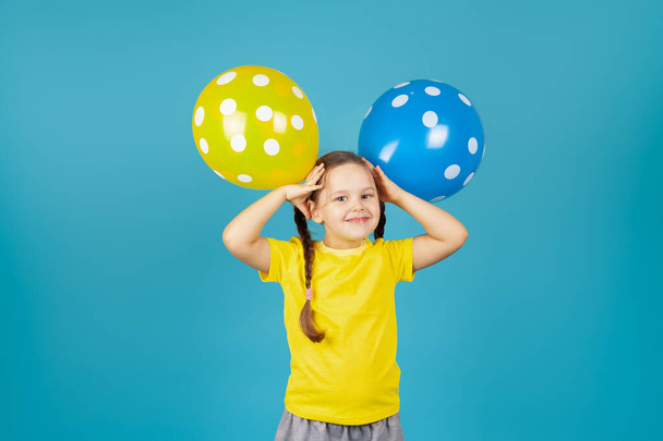 a joyful pigtailed girl in a yellow T-shirt holds balloons at her head like ears, isolated on a blue background - Photo, image