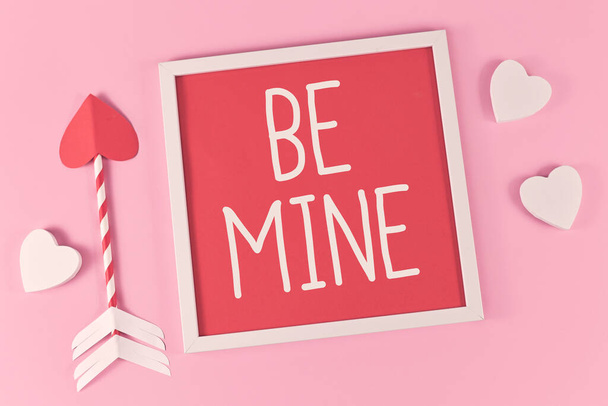 Valentine's Day composition with picture frame with text 'Be Mine', cupid love arrows, and white hearts on pink background - Fotografie, Obrázek