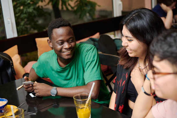 Egy fiatal fekete férfi viccet mesél a barátnőjének, körülvéve barátokkal egy modern kávézóban. - Fotó, kép