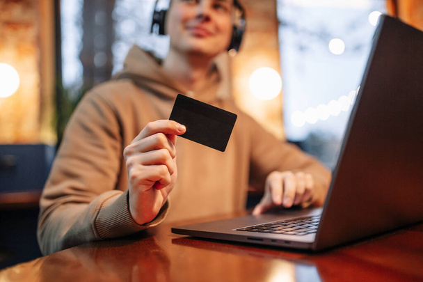 Zakupy i płatności online za pomocą laptopa i karty kredytowej. Mężczyzna siedzący w kawiarni w luźnych ciuchach kupuje towary i płaci przez internet czarną kartą debetową. Nowa koncepcja normalności i technologii - Zdjęcie, obraz