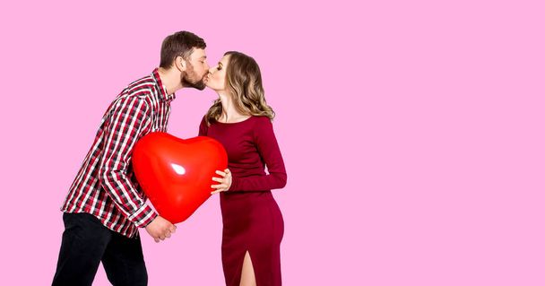 Chica y chico se besan sobre un fondo rosa y sostienen en sus manos una gran bola roja inflable en forma de corazón. Concepto de la fiesta - Día de San Valentín, Día Internacional de la Mujer, Día de la Madre - Foto, imagen
