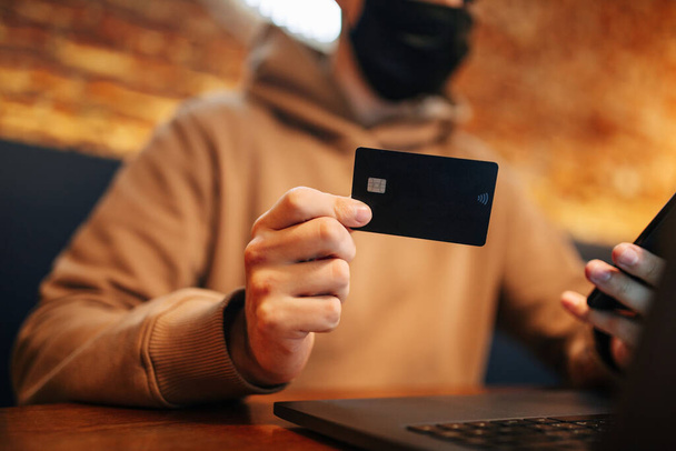 Nakupování a on-line platby pomocí mobilu, notebooku a kreditní karty. Muž v černé lékařské masce během Covid-19 sedí v kavárně a nakupuje zboží přes internet. Nový normální a technologický koncept - Fotografie, Obrázek