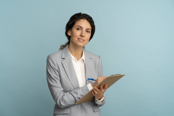 Friendly insurance agent woman wear grey blazer and white blouse holding clipboard, pen looking at camera - Фото, изображение