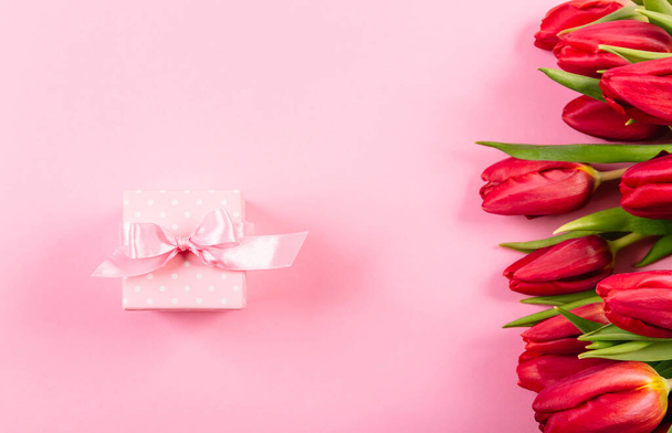 Geschenkdoos met een roze lint in witte stippen op een achtergrond van een boeket rode tulpen met kopieerruimte voor tekst. Behang of banner voor cadeauwinkel, bloemenwinkel of juwelier. Valentijnsdag. - Foto, afbeelding