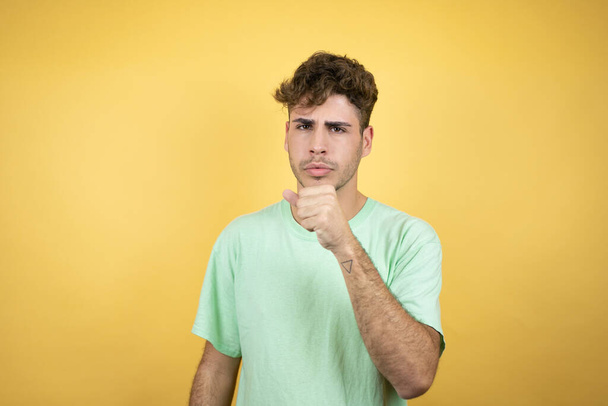 Handsome man wearing a green casual t-shirt over yellow background with his hand to his mouth because he's coughing - Photo, Image