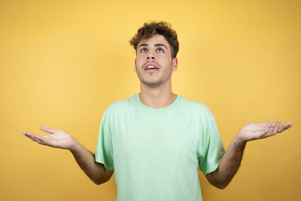 Handsome man wearing a green casual t-shirt over yellow background clueless and confused expression with arms and hands raised - Photo, Image