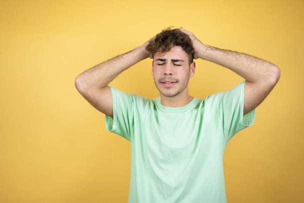 Красивый мужчина в зеленой повседневной футболке на желтом фоне страдает от головной боли отчаянно и стресс, потому что боль и мигрень с руками на голове - Фото, изображение