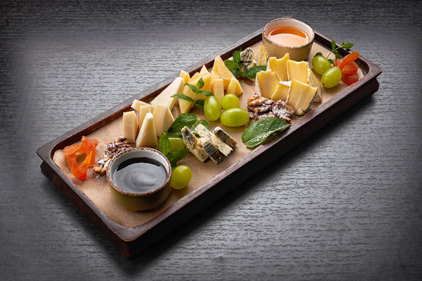 Bandeja de queso sobre una tabla de madera con uvas, menta, miel y mermelada, sobre un fondo oscuro - Foto, Imagen