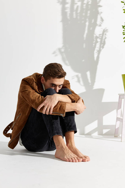 Un tipo con ropa de moda se sienta en el suelo abrazando sus rodillas con las manos En una habitación luminosa - Foto, imagen