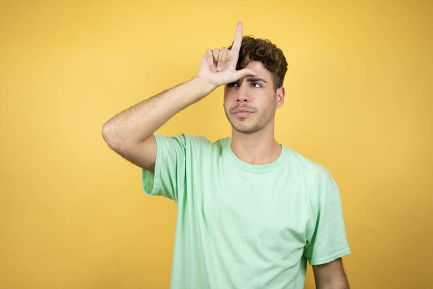 Όμορφος άντρας που φοράει ένα πράσινο casual t-shirt πάνω από κίτρινο φόντο κοροϊδεύοντας ανθρώπους με δάχτυλα στο μέτωπο κάνοντας αποτυχημένες χειρονομίες χλευάζοντας και προσβάλλοντας. - Φωτογραφία, εικόνα
