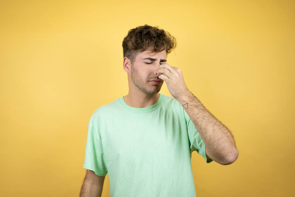 Handsome man wearing a green casual t-shirt over yellow background smelling something stinky and disgusting, intolerable smell, holding breath with fingers on nose - Photo, Image