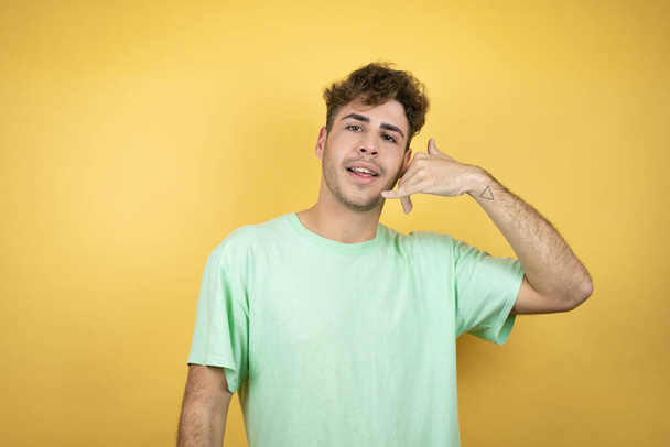 Handsome man wearing a green casual t-shirt over yellow background smiling doing phone gesture with hand and fingers like talking on the telephone - Photo, Image