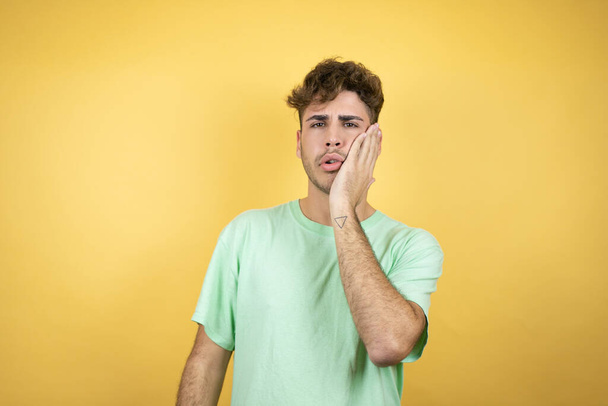歯の痛みや歯の病気のために痛みを伴う式で手で口に触れる黄色の背景に緑のカジュアルなTシャツを着てハンサムな男 - 写真・画像