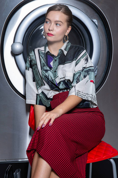 Blond girl model in red skirt sitting with hand near the face washing machine behind - Foto, afbeelding